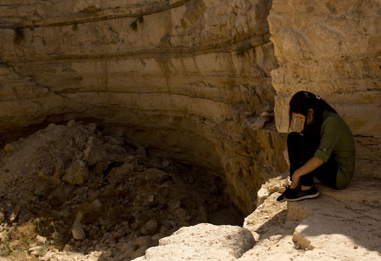 A woman in dark clothes sits, looking forlorn, over a crevice with rubble in it.