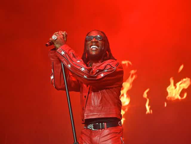 A rebstage backdrop with flames and a man holding a microphone in front of it. He beams, wearing shades and a red leather jacket with stars on the sleeves, his hair dreadlocked.