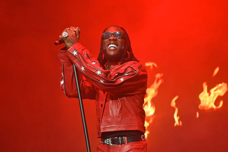 A rebstage backdrop with flames and a man holding a microphone in front of it. He beams, wearing shades and a red leather jacket with stars on the sleeves, his hair dreadlocked.