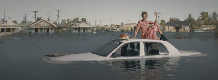 Beyoncé se sienta sobre el techo de un coche de policía cubierto de agua en un pueblo inundado.