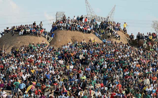 Hundreds of men ands women sit or stand on a hilltop.