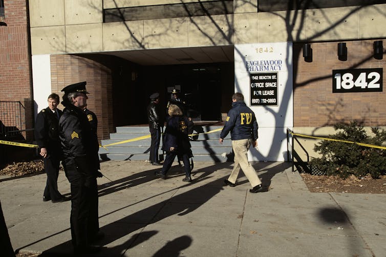Men dressed as police officers and investigators stand outside a generic looking brick building, blocked off with yellow police tape.