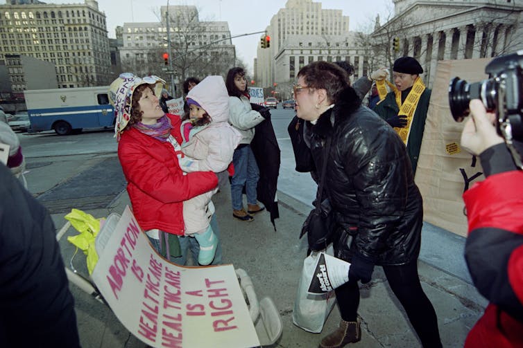 A woman holds a toddler girl in a pink snowsuit and appears to talk in a heated manner with a middle aged woman wearing glasses and a black jacket.