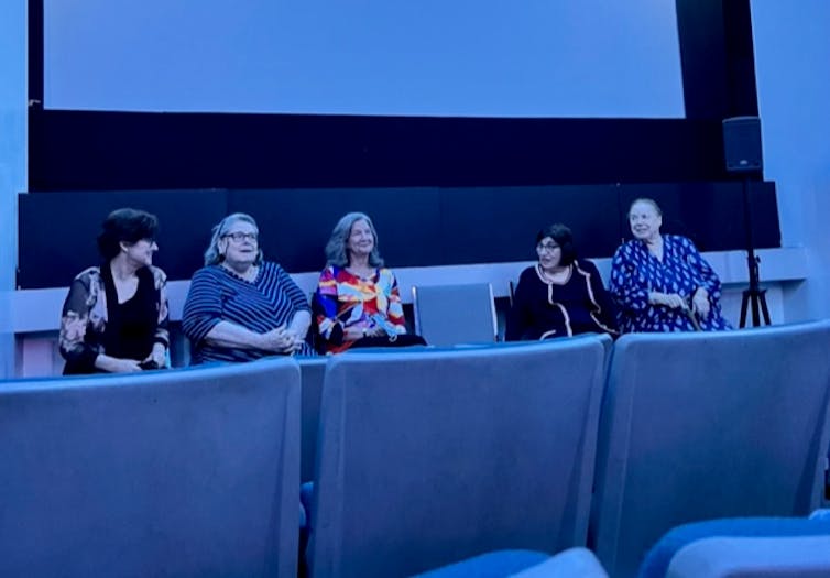 A blue lit room shows five middle aged and older women sitting at the front of an auditorium.