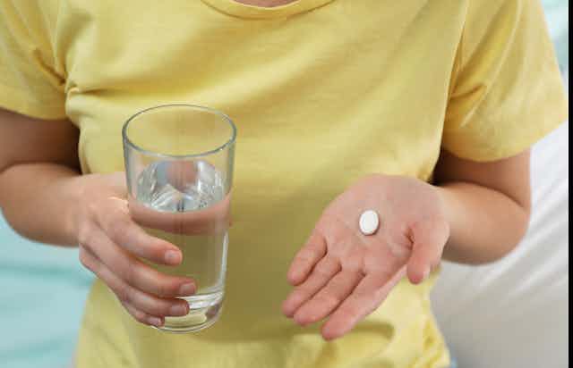 Person wearing a yellow shirt holding a glass of water in one hand and an abortion pill in the other hand.