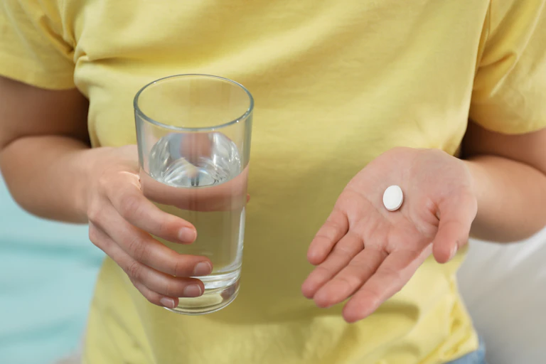 Person wearing a yellow shirt holding a glass of water in one hand and an abortion pill in the other hand.