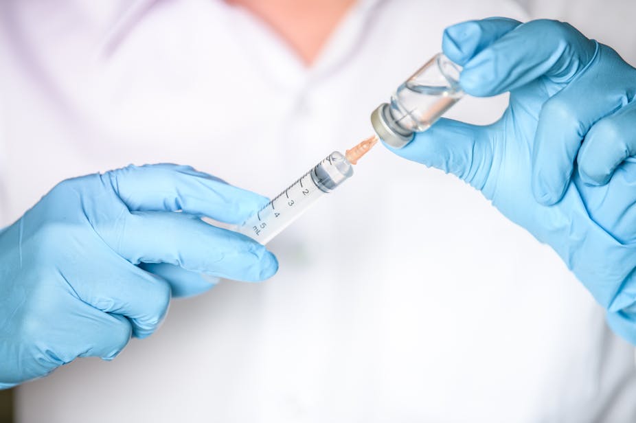 A doctor or nurse wearing blue gloves fills a needle with a vial of clear fluid.