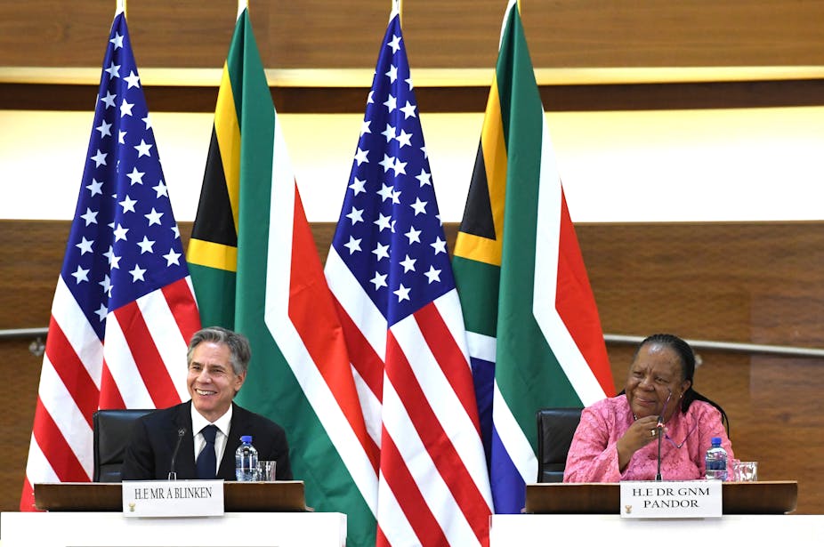 A man wearing a suit and tie, left, and a woman wearing a formal dress smile while sitting behind desks. Behind them are the flags of South Africa and the United States.