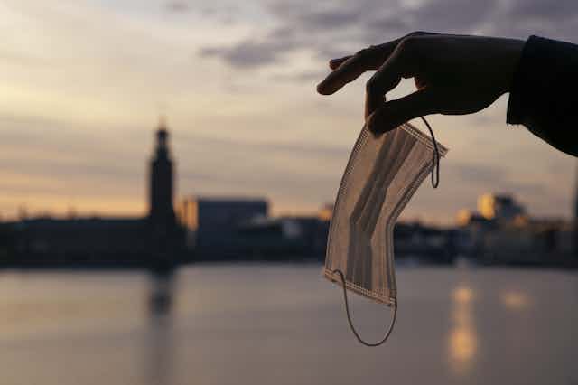 A hand holds a mask in front of Stockholm's skyline.