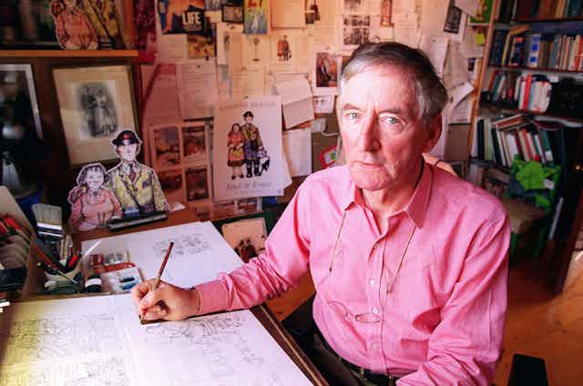 Man in pink shirt at desk surrounded by drawings and books. 
