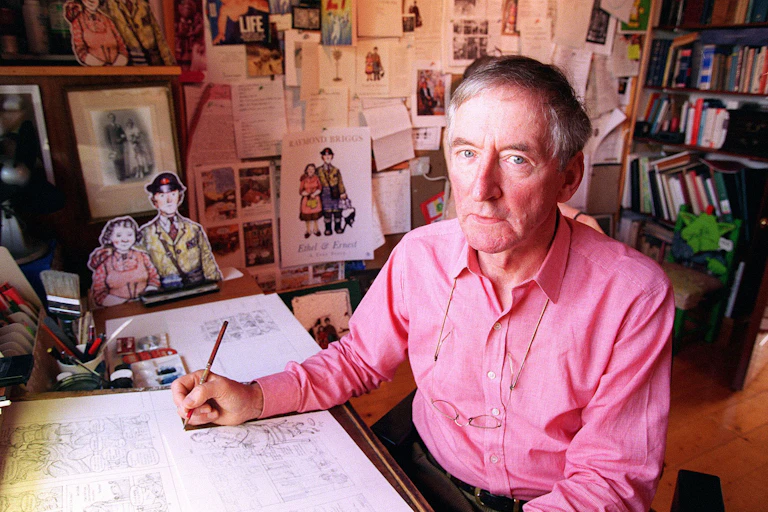 Man in pink shirt at desk surrounded by drawings and books.