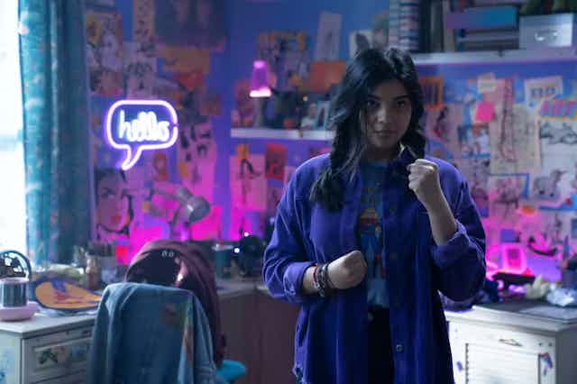 Seorang wanita muda di kamarnya dikelilingi oleh dinding berwarna ungu dan gambar seni dan komik, terlihat mengepalkan satu kepalan di dekat tubuhnya dan mengangkatnya, melihat ke depan.