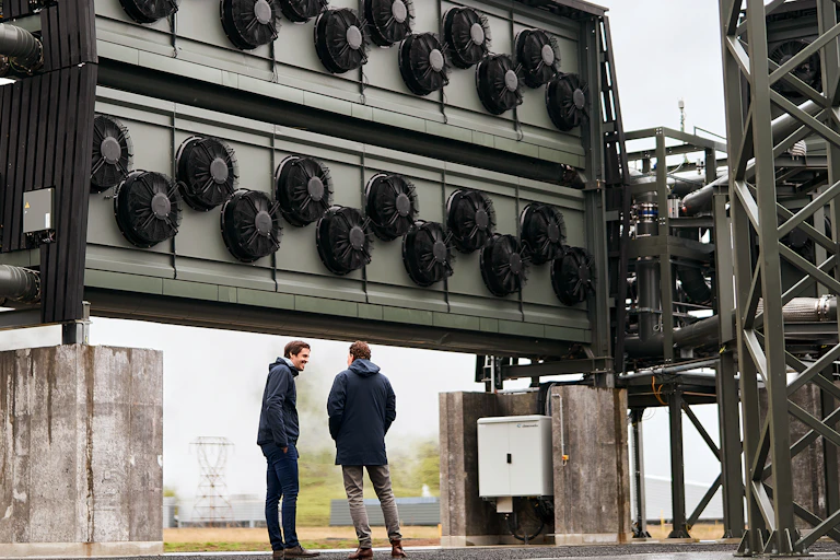 Climeworks Orca plant in Iceland with founders standing in front of a large piece of equipment, about the height of a 2-story house, with fans