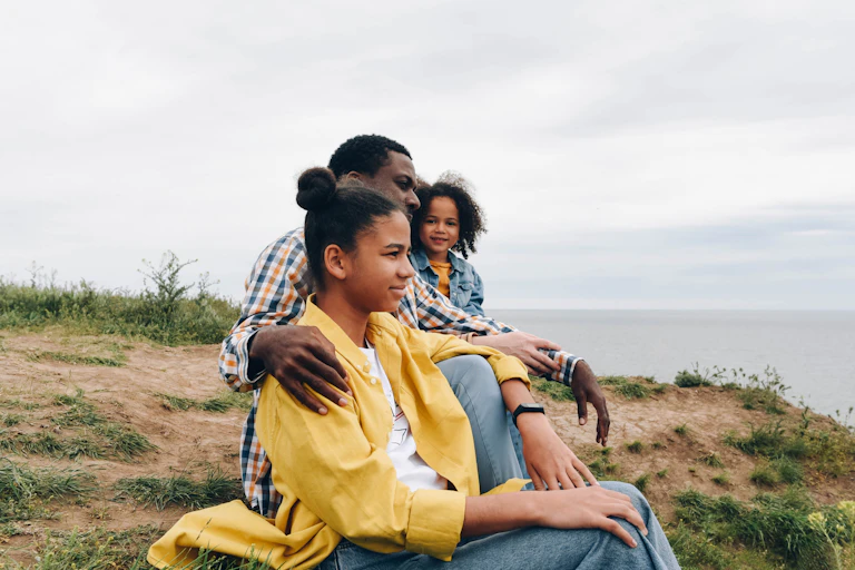A man sits on a hill above the ocean with two young girls sitting on either side of him.