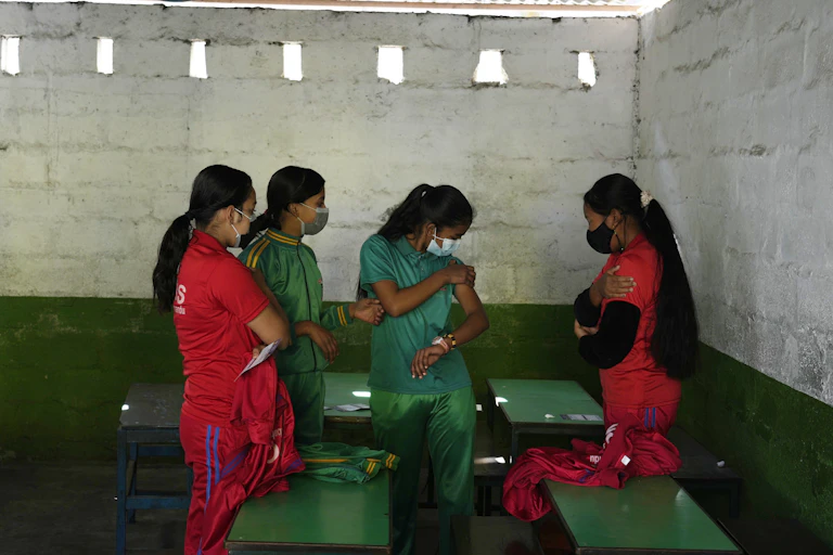 Four girls wearing red and green with face masks on stand in a school house.
