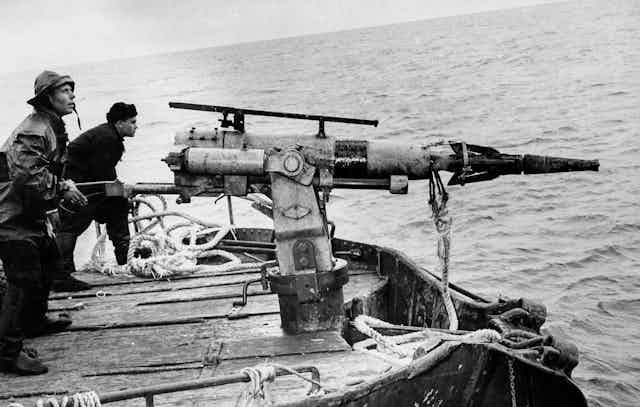 Two men on a ship deck sighting with large spearguns mounted on stands