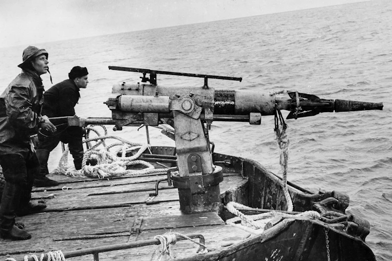 Two men on a ship deck sighting with large spearguns mounted on stands