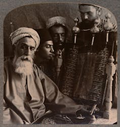 A black and white photo shows four men in turbans looking at a large scroll with text on it.