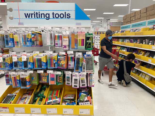 A man wearing a mask stands and gestures toward kneeling son who is looking at school supplies in the writing tools section 
