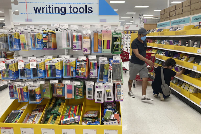 A man wearing a mask stands and gestures toward kneeling son who is looking at school supplies in the writing tools section