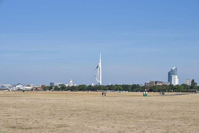A large parched common featuring dead grass with blue sky and a large white tower in the background.