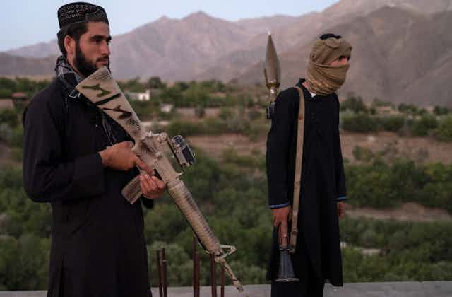 Two Taliban fighters, one wearing a traditional Afghan hat, the other a face covering pose with guns in front of a mountain range.