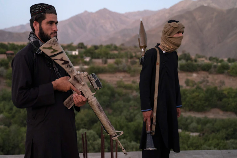 Two Taliban fighters, one wearing a traditional Afghan hat, the other a face covering pose with guns in front of a mountain range.