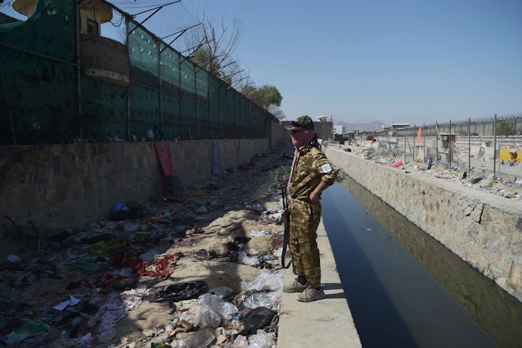 A Taliban fighter holding a gun stands in front of a fence. On the floor is bloodstained clothing and debris.