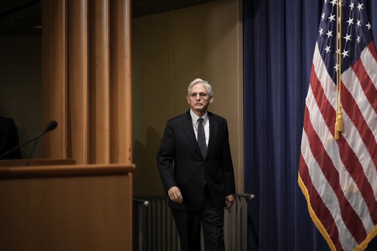 An older white man wearing a black suit is seen walking to a brown lectern, with the American flag standing to his left.