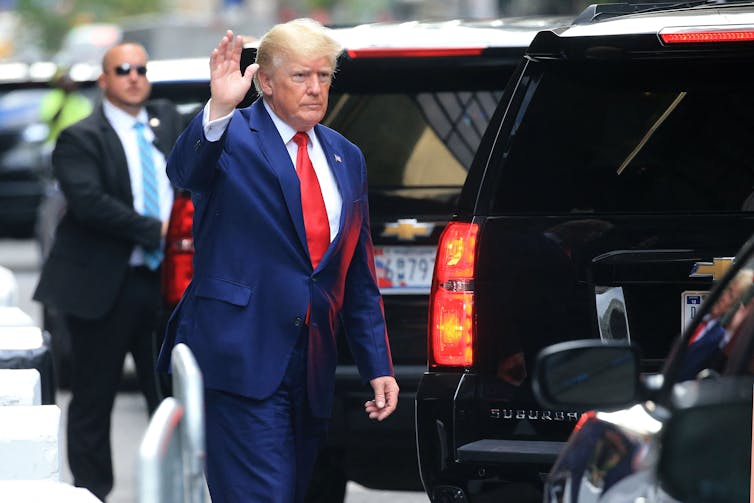 An older white man with white hair, wearing a navy suit and red tie, waves as he walks to a black SUV. Behind him a man wearing sunglasses and a dark suit stands.
