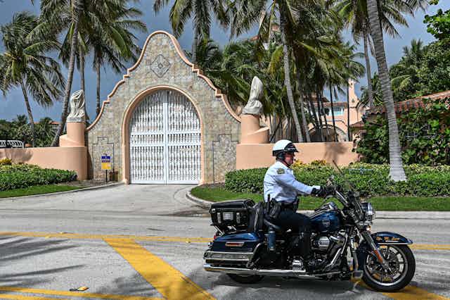 A mn wearing a white jacket and helmet drives a black motorcycle down an empty street, in front of an ornate looking tile and ceramic gate and in front of palm trees.