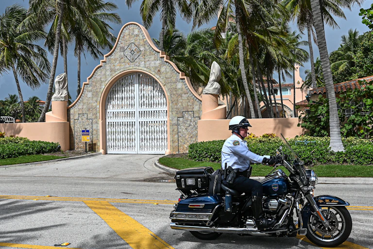 A mn wearing a white jacket and helmet drives a black motorcycle down an empty street, in front of an ornate looking tile and ceramic gate and in front of palm trees.