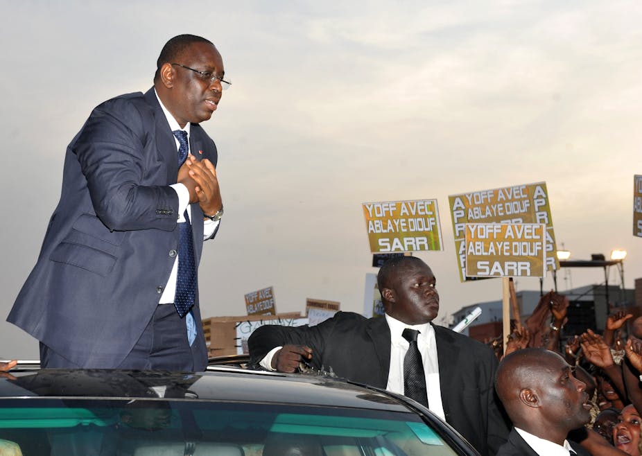 Man in suit stands on a car while greeting a crowd.