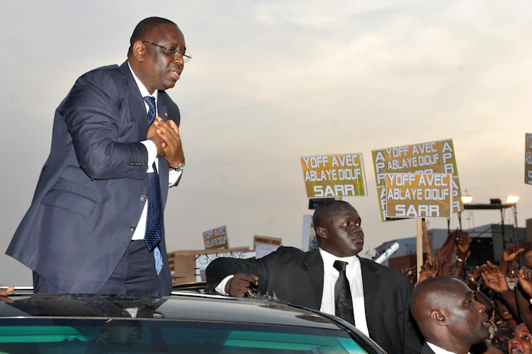 Man in suit stands on a car while greeting a crowd.