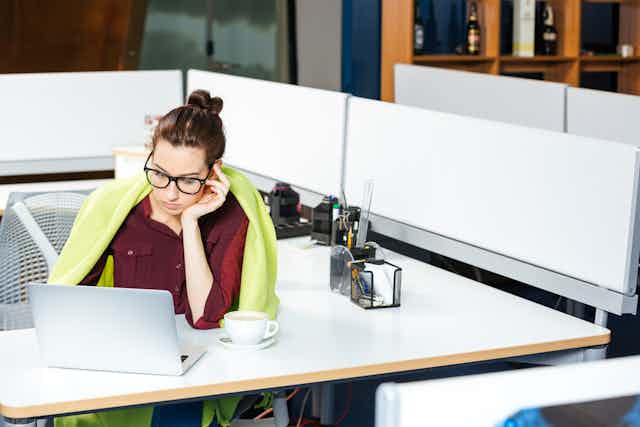 A woman sits at a desk with a blanket around her and a cup of coffee