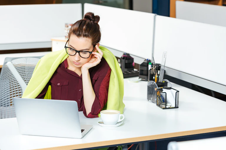 A woman sits at a desk with a blanket around her and a cup of coffee