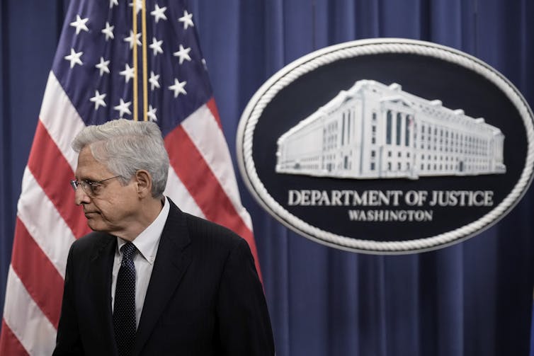 A middle-aged man is leaving a stage as he walks past an American flag and the sign of the U.S. Justice Department.