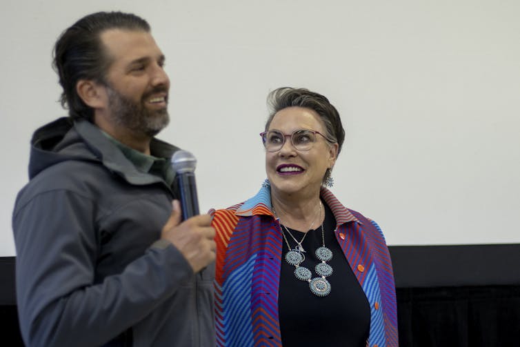 A middle-aged white woman shares a laugh with a bearded man holding a microphone.