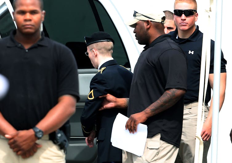 A man in a military uniform is escorted onto a vehicle by a man in a dark shirt and khakis.