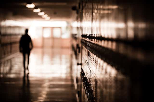 A man walks in a darkened school hallway with lockers on either side. Only his outline can be seen.