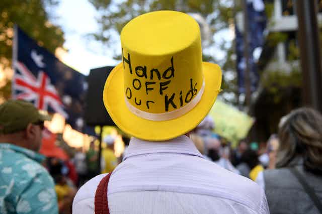 A man wears a large yellow top hat, the back of it reads "hand off our kids"