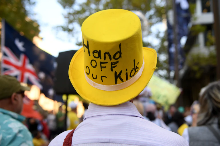 A man wears a large yellow top hat, the back of it reads "hand off our kids"