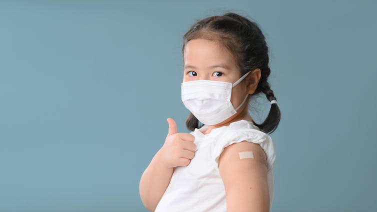 A very young girl with pig-tails is wearing a mask and flashing a thumbs-up to the camera.