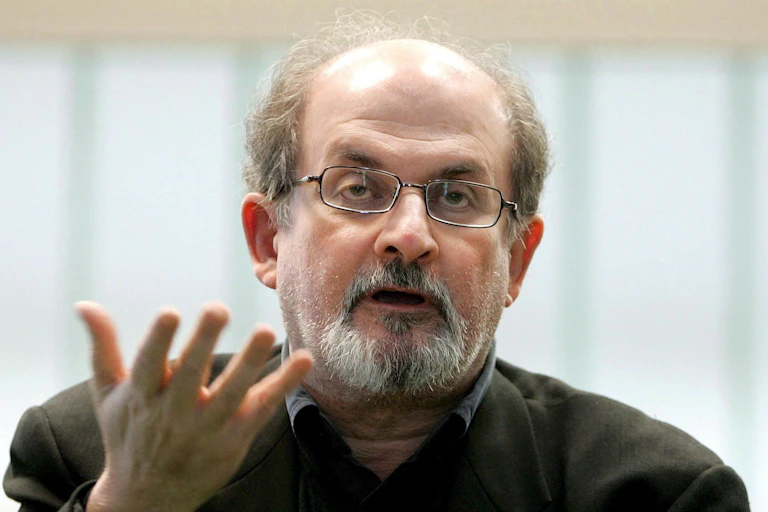 Salman Rushdie, hablando y con una mano extendida.