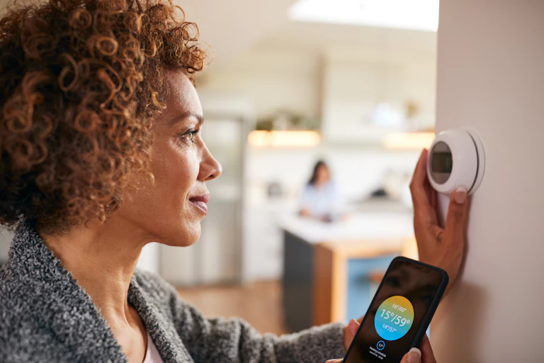 Woman adjusting smart meter while looking at energy bill details on her phone.