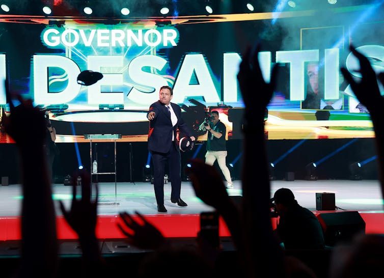 A white man in a blue suit stands on a stage with the words 'Governor De Santis' lit up behind him. He throws a hat into an audience of people.