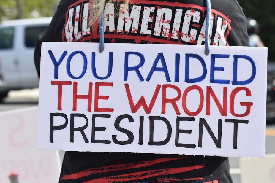 A white sign with blue, red and black writing on someone's back says 'You raided the wrong president.'