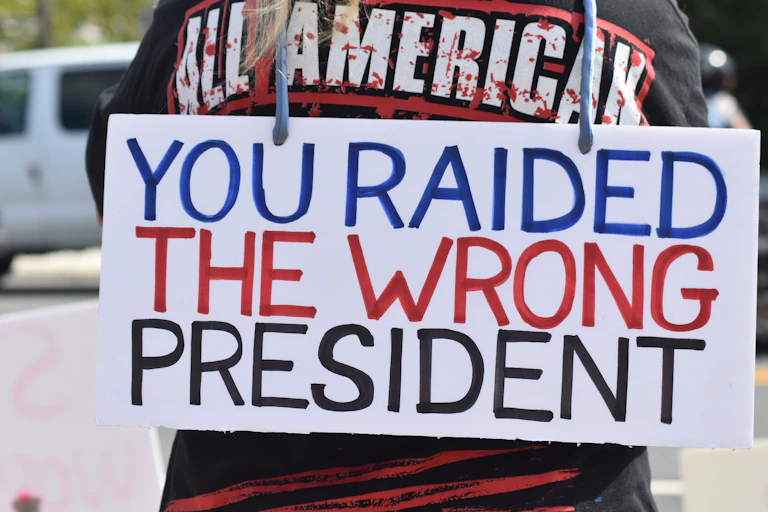 A white sign with blue, red and black writing on someone's back says 'You raided the wrong president.'