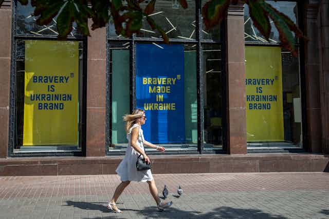A blond white woman wearing a light dress walks by a building with blue and yellow signs that say 'bravery is a Ukrainian brand.'