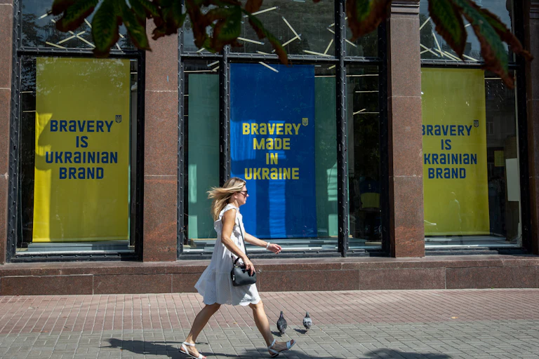 A blond white woman wearing a light dress walks by a building with blue and yellow signs that say 'bravery is a Ukrainian brand.'